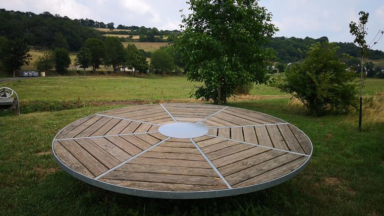Eine runde Holzplattform steht in einer grünen Wiese unter einem Baum. Im Hintergrund sind sanfte Hügel und ein blauer Himmel zu sehen.