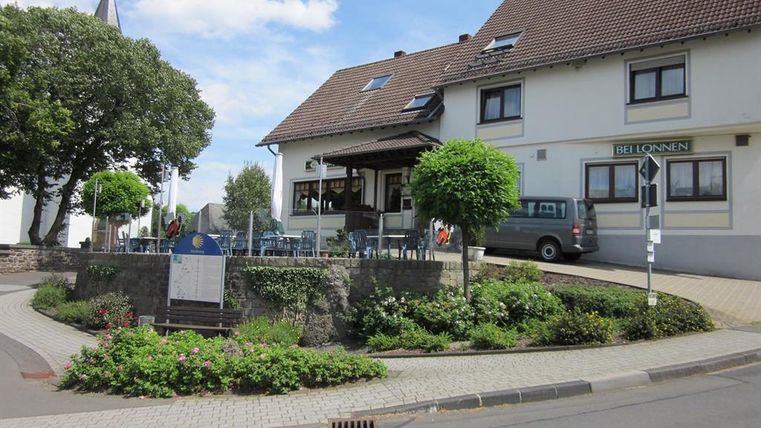 View from the front of the inn Bei Lonnen.
The two-story building is brightly painted and has a black tiled roof.
In front of the guesthouse, you can see a round terrace equipped with several tables and chairs.