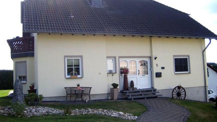 Een gezellig, geel huis met een rood dak en een mooie tuin. Voor het huis staan een tafel en stoelen, evenals decoratieve planten.