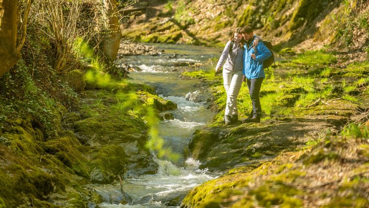 Zwei Personen wandern an einem kleinen, klaren Bach entlang. Die Umgebung ist grün und naturbelassen.