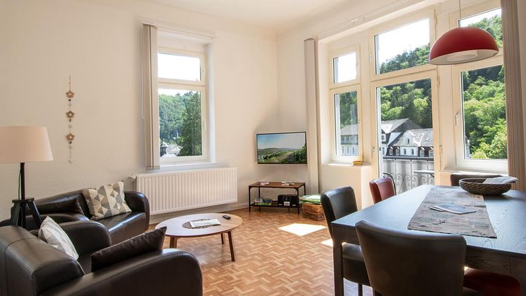 Ein helles Wohnzimmer mit großen Fenstern und Blick ins Grüne. Es gibt bequeme Möbel und einen Esstisch.