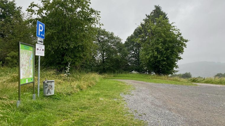 Een parkeerplaats in de natuur, omringd door bomen en gras. De lucht is bewolkt en de weg leidt het groen in.
