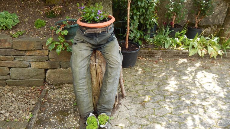 A creative garden decoration depicts a person made of old pants and boots, with a flower pot on their head. Surrounded by plants, it adds a charming touch to the garden.