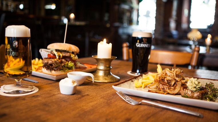 Eine rustikale Tischdekoration mit zwei Tellern Essen, einem Burger und einem Steak, umgeben von Pommes frites. Dazu sind zwei Gläser Bier und eine kerzenbeleuchtete Atmosphäre zu sehen.