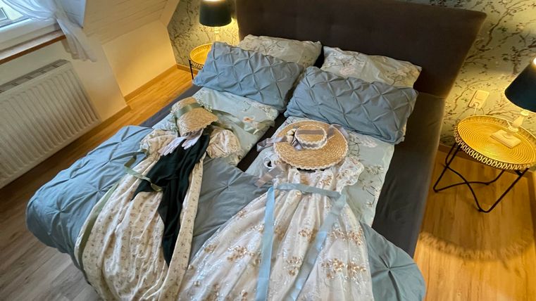 A bedroom with a large bed covered with blue and white bed sheets. Two dresses and a hat are lying on the bed, and the room has a cozy atmosphere.