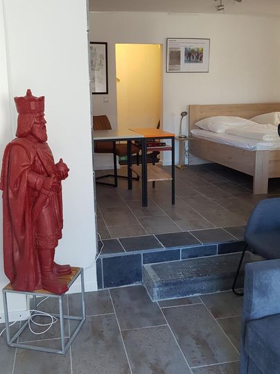 Ein modernes Zimmer mit einem hellen Schlafbereich und einem kleinen Tisch. Eine rote Statue steht in der Ecke, was dem Raum einen kreativen Akzent verleiht.