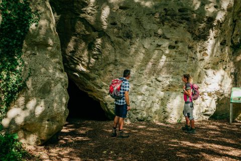 Zwei Wanderer stehen vor einer Höhle, umgeben von Felsen und Bäumen. Die Atmosphäre ist ruhig und naturnah.