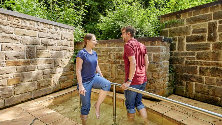 Ein Paar steht in einem flachen Wasserbecken mit Steinmauern und begrüntem Hintergrund. Sie genießen gemeinsam einen entspannenden Moment in der Natur.