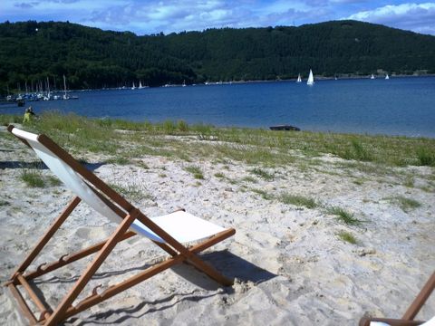 Ein Liegestuhl steht am Ufer eines ruhigen Gewässers. Im Hintergrund sind sanfte Hügel und Segelboote zu sehen.
