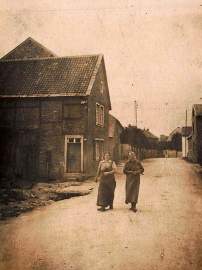 Twee vrouwen lopen door een rustige straat, omringd door oude gebouwen. Het tafereel heeft een nostalgisch karakter en straalt een historische sfeer uit.