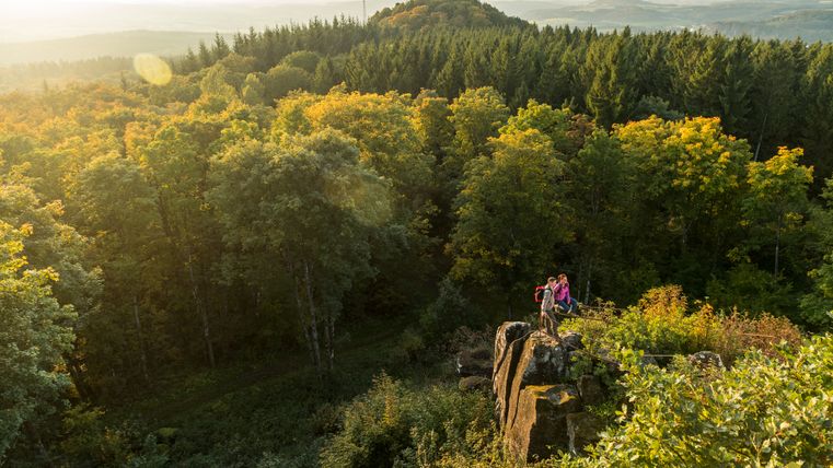 Zwei Personen stehen auf einem Felsen mit Blick auf einen bewaldeten Hügel im Sonnenuntergang.