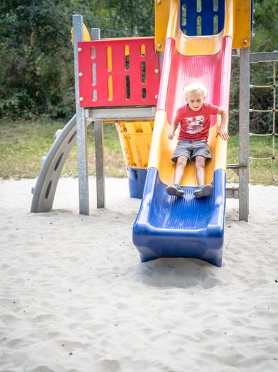 Ein Kind rutscht auf einer bunten Rutsche auf einem Spielplatz. Der Sandboden um die Rutsche sorgt für Sicherheit beim Spielen.