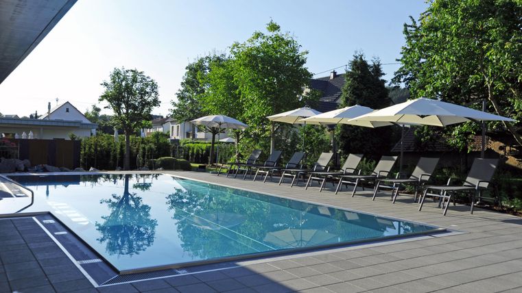 Ein Swimmingpool umgeben von Liegen und Sonnenschirmen. Die Umgebung ist grün mit Bäumen und dem blauen Himmel.