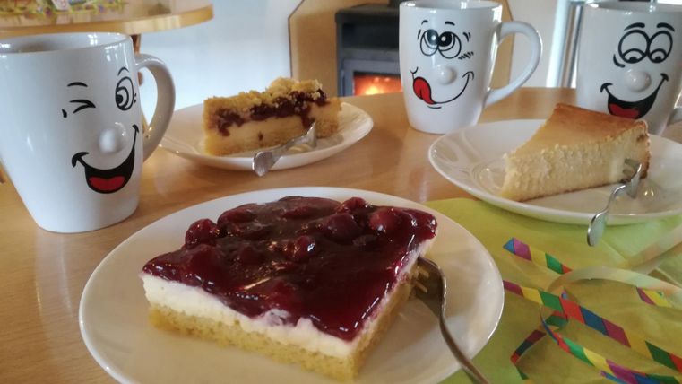 Eine gemütliche Kaffeetafel mit zwei verschiedenen Kuchenstücken und Kaffeetassen mit freundlichen Gesichtern. Im Hintergrund sieht man einen Kamin, der eine warme Atmosphäre schafft.