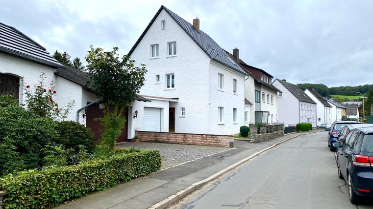 Een stille straat met witte woonhuizen en een licht bewolkte lucht. Aan de linkerkant bevinden zich kleine tuinen en geparkeerde auto's.