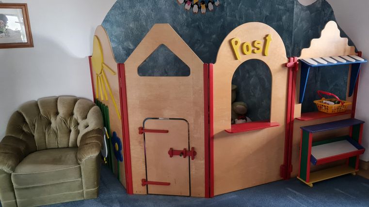 Ein bunter Spielbereich mit einer kleinen Holzhütte und einem Spieltisch. Neben einem gemütlichen Sessel steht die Hütte mit einem Postfenster.