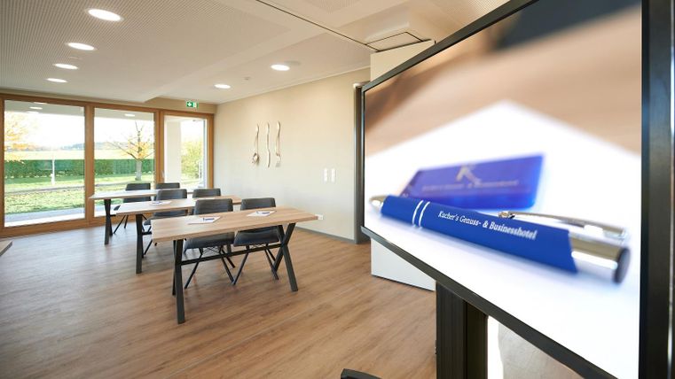 Ein moderner Konferenzraum mit Tischen und Stühlen. Ein Bildschirm zeigt einen nahen Blick auf einen blauen Stift.