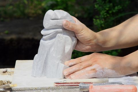 A person is working on a sculpture made of stone. The hands are holding a part of the unfinished sculpture, while tools are lying on the table.