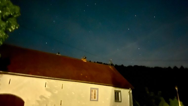Ein altes Haus bei Nacht, beleuchtet von Sternen am Himmel. Die Umgebung ist dunkel und ruhig, mit Bäumen im Hintergrund.