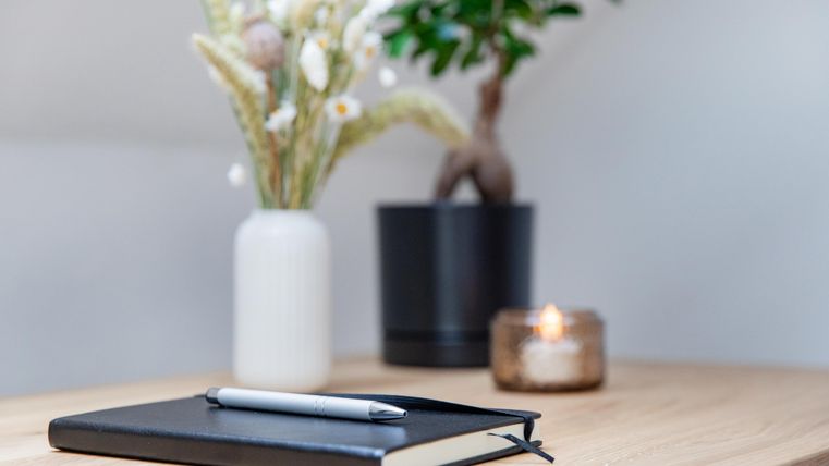 Ein schwarzes Notizbuch liegt auf einem Holztisch mit einem Stift darauf. Im Hintergrund sind eine Vase mit Blumen, eine Pflanze und eine Kerze zu sehen.
