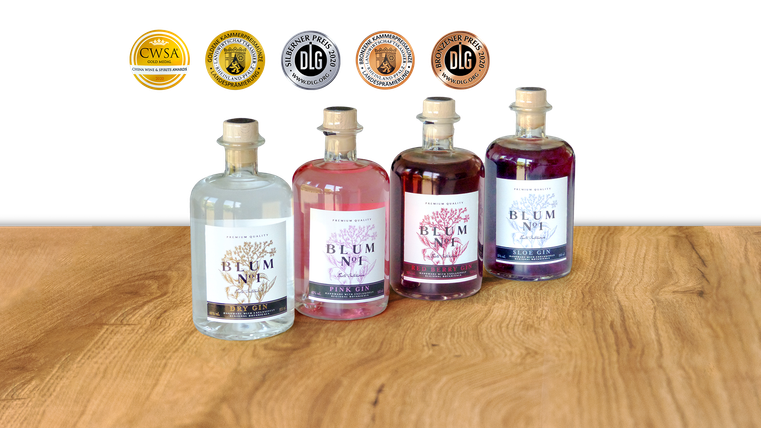 Vier Flaschen Blum Gin in verschiedenen Sorten stehen auf einem Holztisch. Im Hintergrund sind mehrere Auszeichnungen zu sehen.