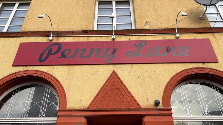 Een rood geborduurd winkeltje met de naam "Penny Lane". De gevel is lichtgeel met grote ramen.