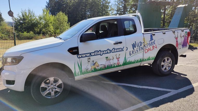 A white pickup with a colorful sticker advertising the wildlife park. In the background, there are trees and a blue sky.