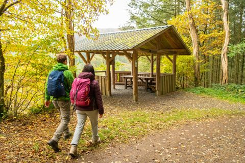 Zwei Wanderer gehen auf einem Weg zu einer Überdachung im Wald. Die Bäume zeigen bunte Herbstblätter.