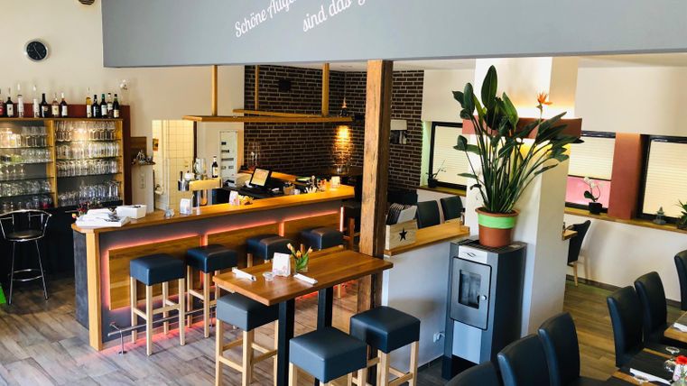 Modern restaurant with bar, wooden tables and chairs. On the wall, it says "Beautiful moments are the happiness of life." Plants and glasses decorate the room.