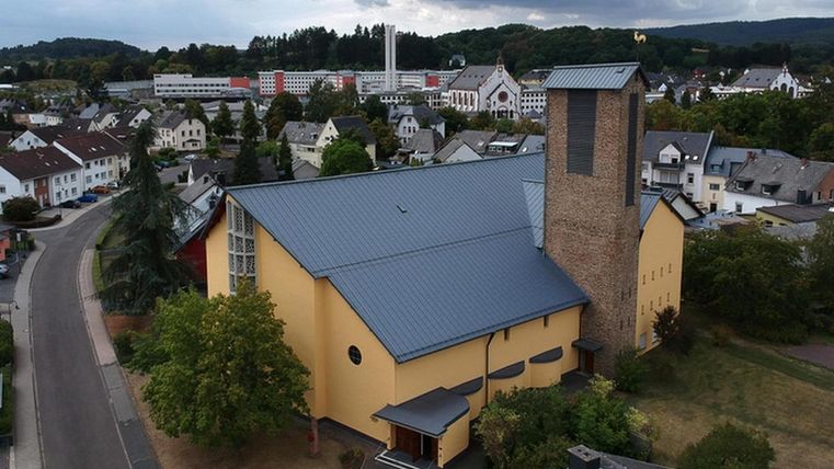 Een moderne kerk met een gele gevel en een hoge toren. Op de achtergrond is een stadslandschap met meerdere gebouwen en heuvels zichtbaar.