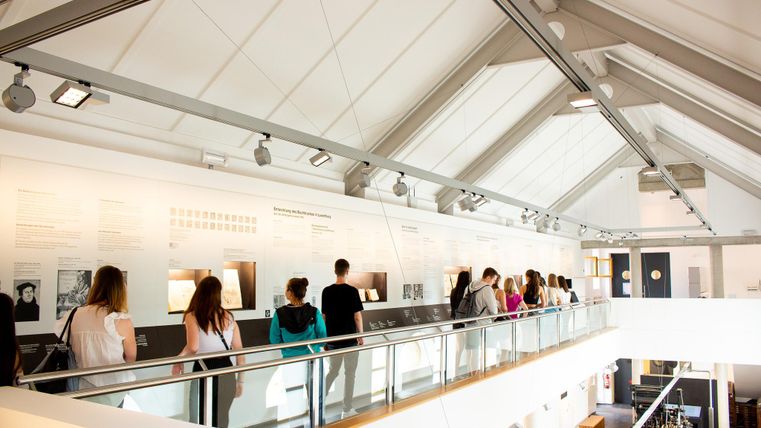 A modern exhibition space with long walls displaying images and information. Visitors stand in a row and observe the exhibit.