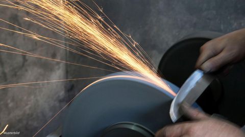 Een mes wordt geslepen op een slijpsteen, terwijl vonken overspringen. Handen houden het mes stevig vast.