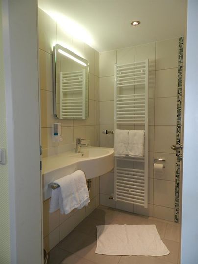 Een moderne badkamer met lichte tegels en een wastafel. Handdoeken zijn netjes geplaatst, en een grote spiegel geeft de ruimte een ruimtelijk gevoel.