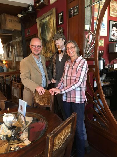 Ein Mann und eine Frau stehen lächelnd neben einer Sherlock-Holmes-Figur in einem gemütlich dekorierten Raum mit Holzmöbeln und Bildern.