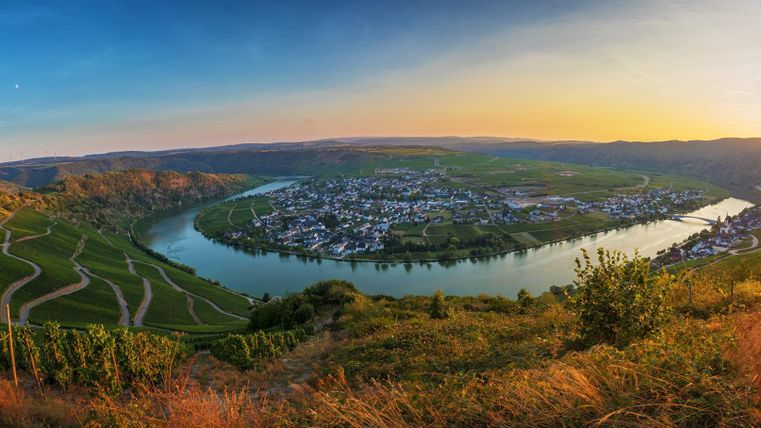 Eine schöne Panoramaansicht des Flusses mit Weinbergen im Vordergrund. Der Himmel leuchtet in warmen Farben während des Sonnenuntergangs.