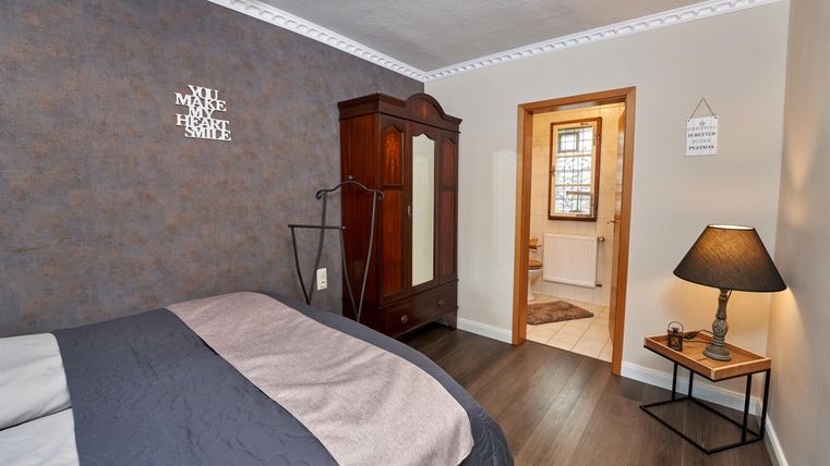 A cozy bedroom with a large bed and a classic wardrobe. A nightstand with a lamp and access to another room are also visible.