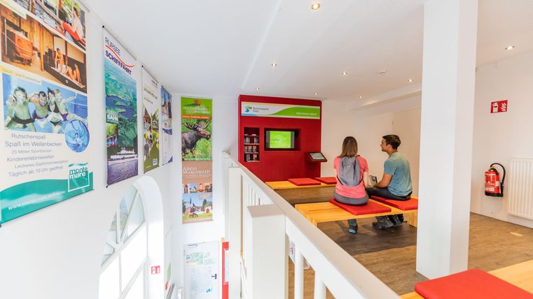 Een modern informatiecentrum met kleurrijke posters aan de muren. Twee personen zitten en kijken naar een scherm.