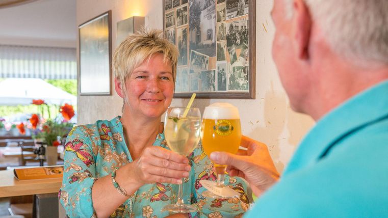 Eine Frau prostet mit einem Getränk einem Mann zu. Im Hintergrund sind gedeckte Tische und alte Fotos zu sehen.