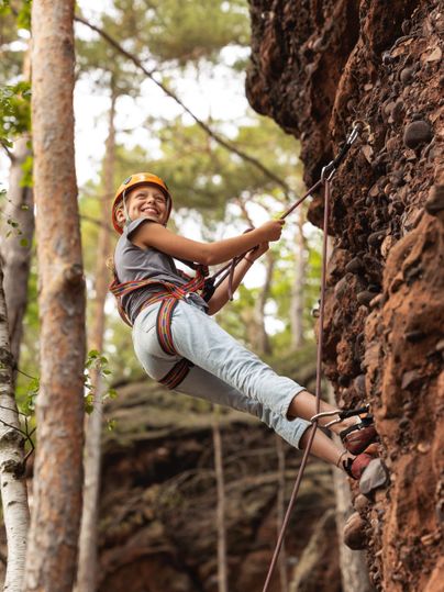 Een vrolijk kind in een klimmuts klimt aan een rotswand. Omringd door bomen geniet het van het avontuur in de natuur.