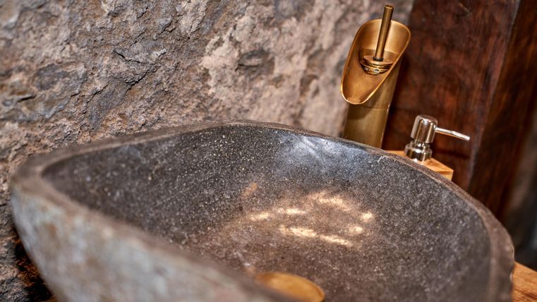 An elegant sink made of dark stone with golden fixtures. The rustic surroundings of stones and wood complement the modern design.
