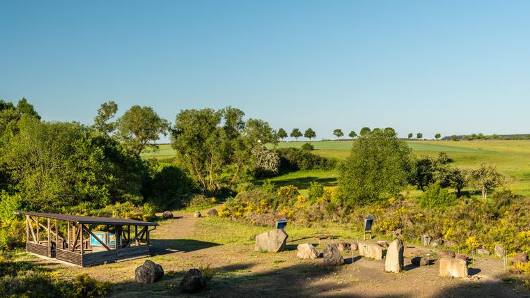 Eine ruhige Landschaft mit grünen Wiesen und Bäumen. Im Vordergrund befinden sich einige Steine und eine überdachte Sitzgelegenheit.