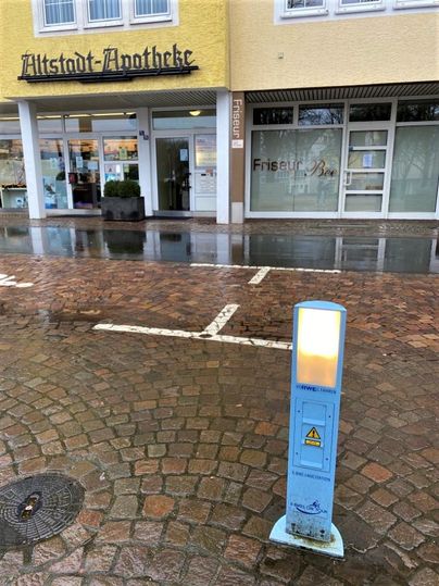 A pharmacy with a yellow facade and a hair salon next to it. In the foreground, there is a blue traffic light.