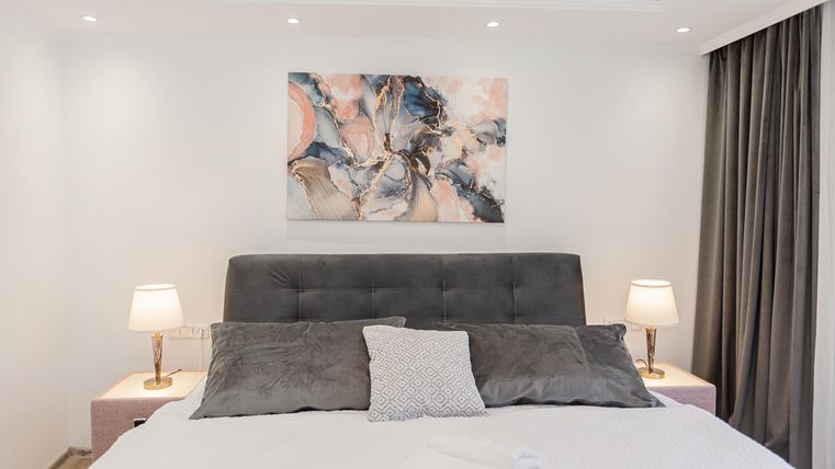 Ein elegantes Schlafzimmer mit einem grauen Bett und sanften Kissen. An der Wand hängt ein modernes Kunstwerk, und zwei Nachtlichter sorgen für eine gemütliche Atmosphäre.