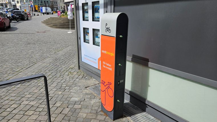 Eine Fahrradstation mit einem orangefarbenen Informationsschild steht auf einem Pflasterweg. Im Hintergrund sind mehrere Gebäude und parkende Autos zu sehen.