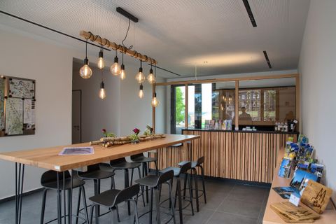 Ein modernes Foyer mit einem langen Holztisch und stilvollen Stühlen. An der Wand befinden sich Informationsmaterialien und große Fenster sorgen für viel Tageslicht.