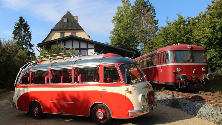 Ein alter, roter und cremefarbener Reisebus steht neben einer roten Lokomotive auf einem Gleis. Im Hintergrund ist ein Haus mit einem spitzen Dach zu sehen.