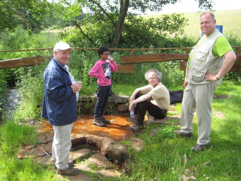 <p>Drei Männer und ein Kind stehen um eine kleine viereckige Umrandung im Boden , aus der Wasser abfliesst. Der Drees ist mit Platten umlegt und   umrahmt von einer Wiese.</p>