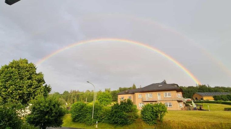 Ein Regenbogen spannt sich über ein Grundstück mit einem Haus und viel Grün. Der Himmel ist grau mit einigen Wolken.