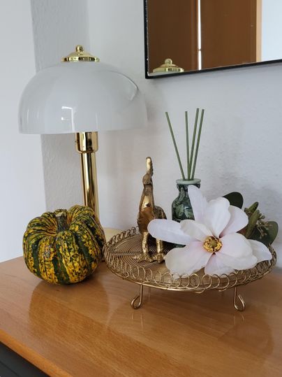 A decorative tabletop arrangement with a lamp, a golden figure, and floral decoration. On the table lies a woven basket with a pumpkin and other decorative elements.