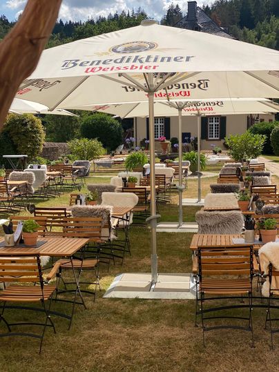 Een gezellige buitenruimte met houten stoelen en tafels, die onder grote parasols staan. De tuin is groen en uitnodigend, ideaal voor een ontspannen tijd buiten.